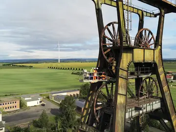 Eine Luftbildaufnahme eines Fördergerüstes mit landschaftlichem Hintergrund. Mittels Hubsteiger wird eine Prüfung durchgeführt.